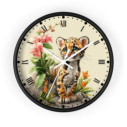 Round wall clock with a black frame and a cream-colored face featuring an illustrated Ocelot cat, pink flowers, green leaves, and small orange blossoms. Roman numerals mark the hours, and the clock hands are white.