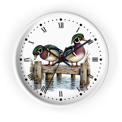 Round wall clock with a white face featuring an illustration of two colorful wood ducks standing on a wooden dock over water, with black Roman numerals and hands on a white background.