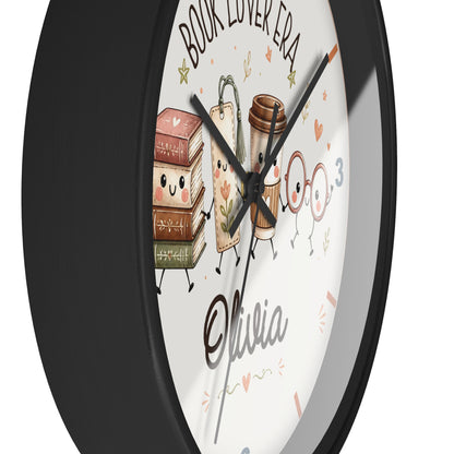 A round wall clock with a black frame and a white face featuring cute illustrations of books, a coffee cup, glasses, and the text 'BOOK LOVER ERA' and 'Olivia'.