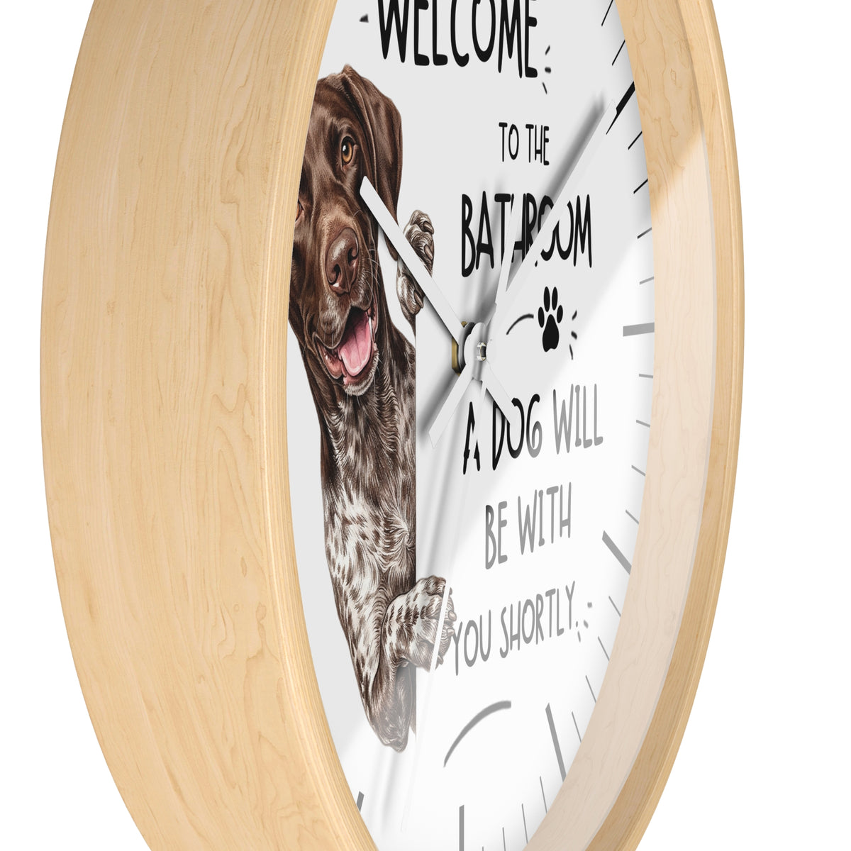 A round wall clock with a light natural wood frame and a white face. The clock face features a printed brown dog illustration and the humorous text 'WELCOME TO THE BATHROOM A DOG WILL BE WITH YOU SHORTLY'.
