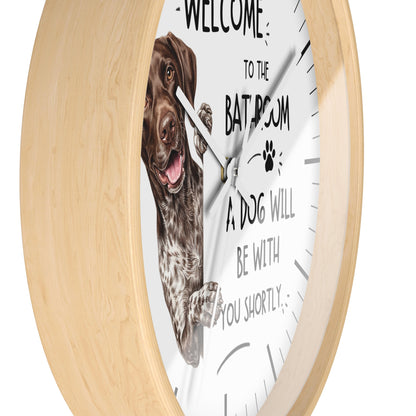 A round wall clock with a light natural wood frame and a white face. The clock face features a printed brown dog illustration and the humorous text 'WELCOME TO THE BATHROOM A DOG WILL BE WITH YOU SHORTLY'.