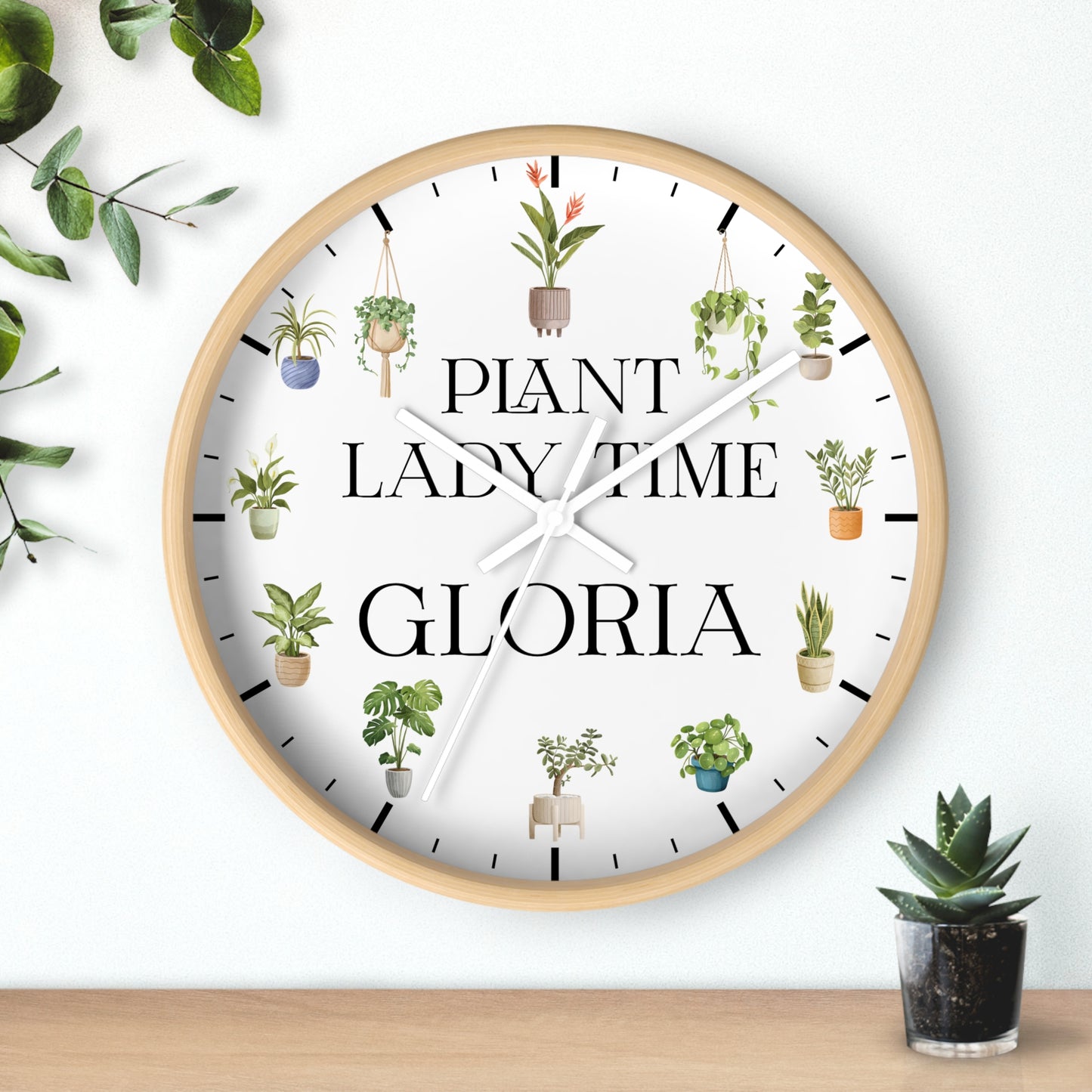 A round wall clock with a light wood frame and a white face, featuring the text 'PLANT LADY TIME GLORIA' and small illustrated potted plants around the dial. The clock is mounted on a white wall above a wooden surface, with decorative green leaves and a small succulent nearby.