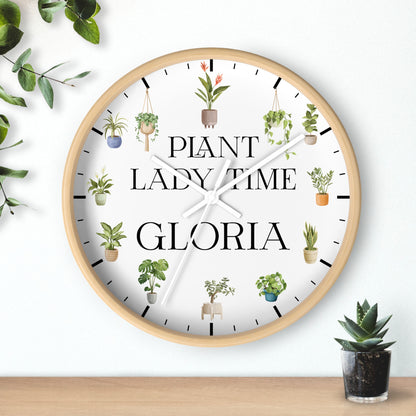 A round wall clock with a light wood frame and a white face, featuring the text 'PLANT LADY TIME GLORIA' and small illustrated potted plants around the dial. The clock is mounted on a white wall above a wooden surface, with decorative green leaves and a small succulent nearby.