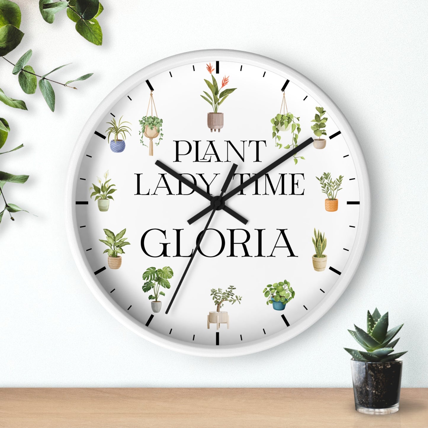 A round white wall clock with black hands and black hour markers, decorated with illustrations of potted plants and the text 'PLANT LADY TIME GLORIA'. The clock is shown hanging on a white wall above a wooden surface with small potted plants nearby.