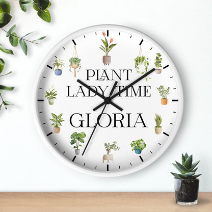 A round white wall clock with black hands and black hour markers, decorated with illustrations of potted plants and the text 'PLANT LADY TIME GLORIA'. The clock is shown hanging on a white wall above a wooden surface with small potted plants nearby.