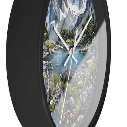 A round wall clock with a black frame and a scenic landscape print featuring a mountain lake, trees, and wildflowers. The clock is shown at an angle against a plain white background.