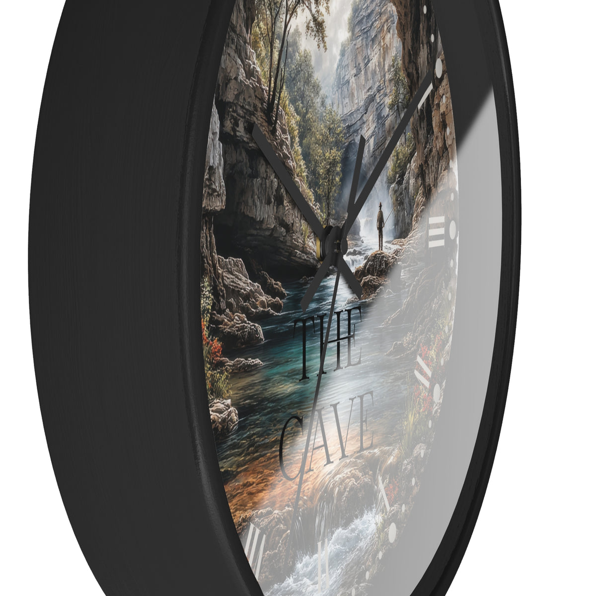 A round wall clock with a black frame and a scenic landscape print inside showing a cave stream, trees, and the words “THE CAVE,” photographed against a white background.