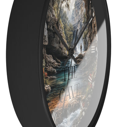 A round wall clock with a black frame and a scenic landscape print inside showing a cave stream, trees, and the words “THE CAVE,” photographed against a white background.