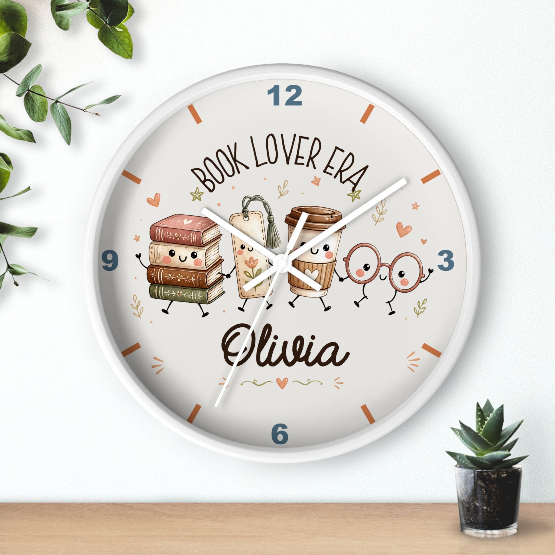 A personalized round wall clock with a white frame, featuring cartoon illustrations of books, a coffee cup, and reading glasses with the text 'BOOK LOVER ERA' and the name 'Olivia'. The clock is styled on a light wall above a wooden surface with small green plants.