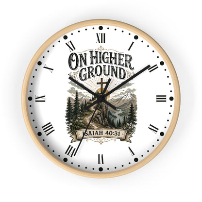 A round wall clock with a light natural wood frame, white face, black Roman numerals, and a scenic graphic featuring mountains, pine trees, and a cross. The text on the clock reads 'ON HIGHER GROUND' and 'SAITH 40:31'.