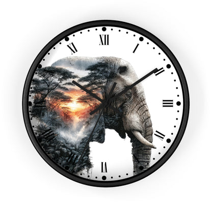 A round wall clock with a black frame and a white face featuring a large elephant silhouette filled with an African savanna landscape scene, including trees, mist, and a warm sunset. The clock has black Roman numerals and black hands on a plain white background.