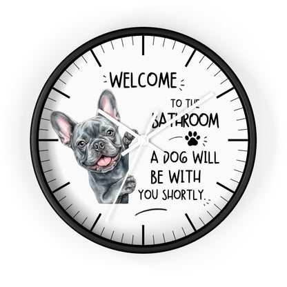 A round wall clock with a black frame and a white face, featuring a French bulldog illustration and the text 'WELCOME TO THE BATHROOM A DOG WILL BE WITH YOU SHORTLY.' on a light gray background.