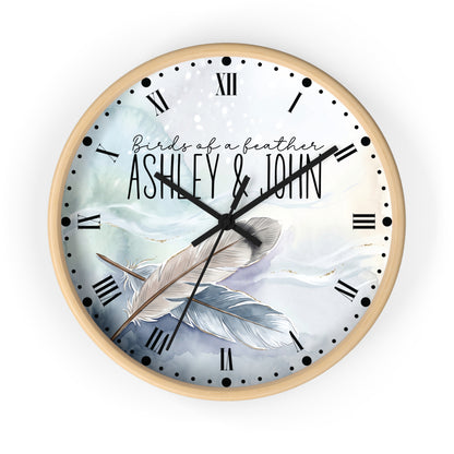 A round wall clock with a light wood frame and a printed dial featuring the text 'Birds of a feather ASHLEY & JOHN' over an illustration of two feathers on a soft blue and white background.