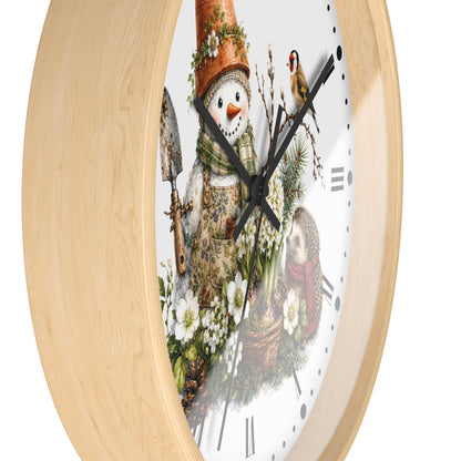 A round wall clock with a light natural wood frame and a white face featuring a whimsical winter garden illustration. The design shows a snowman wearing a terracotta flower pot hat and green scarf, holding a trowel, with a small bird perched on the clock hands and Roman numeral hour markers.