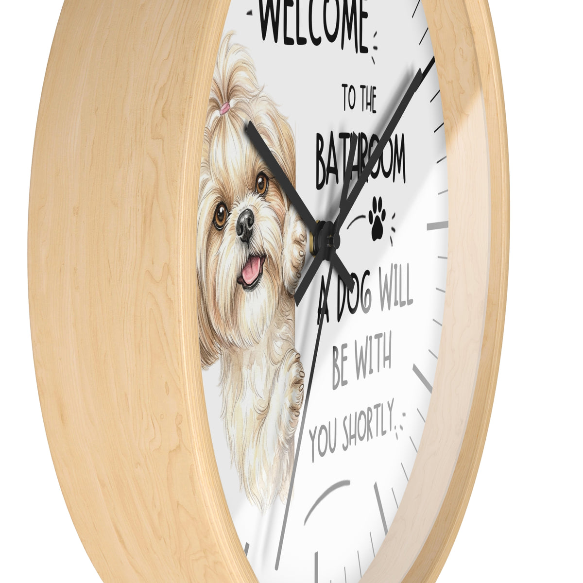 A round wall clock with a light natural wood frame and a white face featuring an illustration of a small fluffy dog and the text 'WELCOME TO THE BATHROOM A DOG WILL BE WITH YOU SHORTLY'.