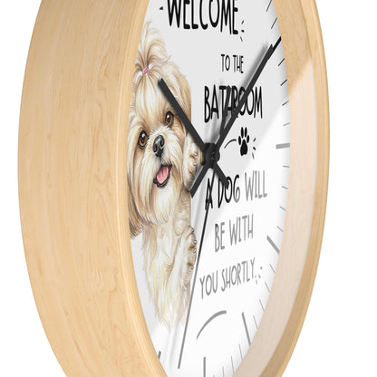 A round wall clock with a light natural wood frame and a white face featuring an illustration of a small fluffy dog and the text 'WELCOME TO THE BATHROOM A DOG WILL BE WITH YOU SHORTLY'.