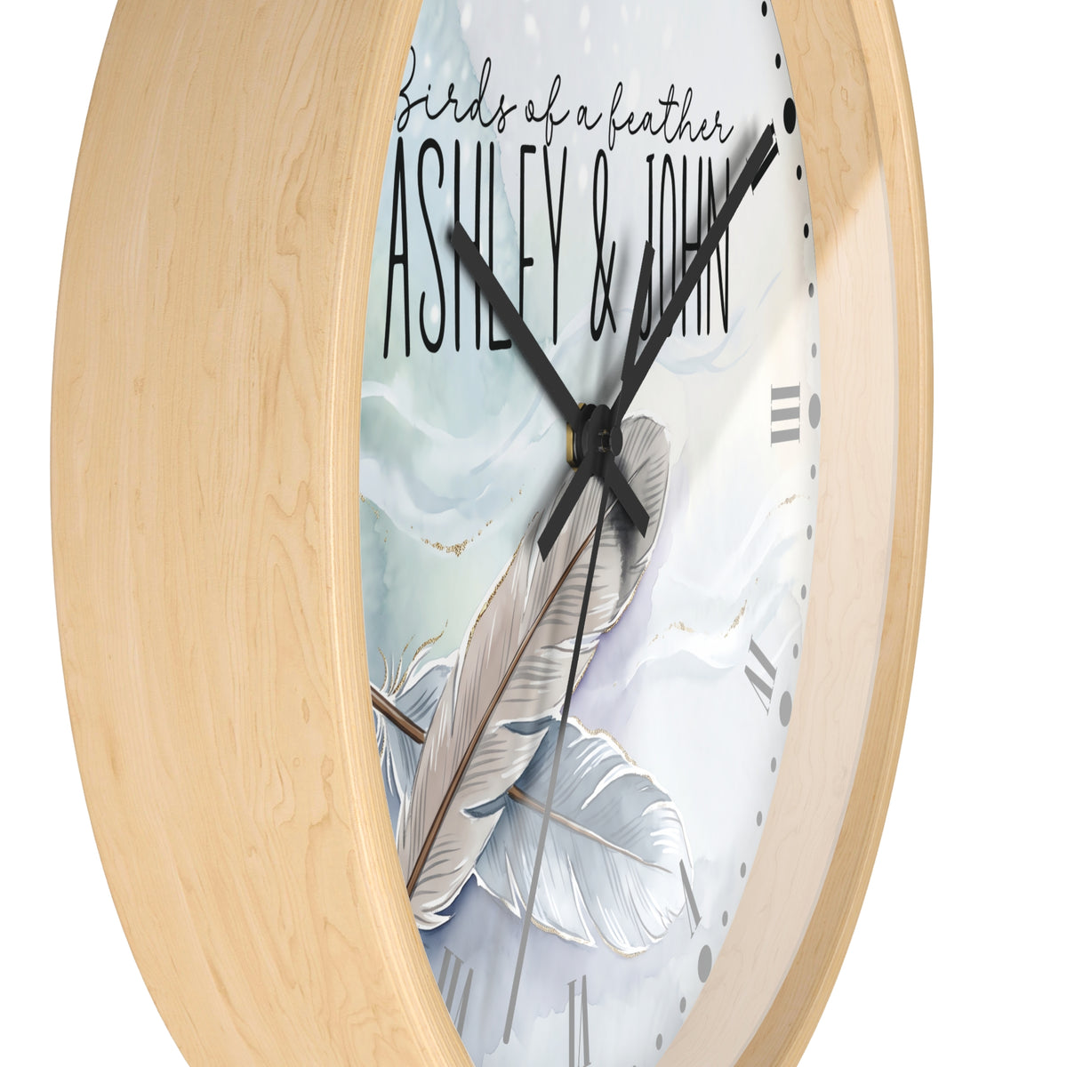 Oval wall clock with a light natural wood frame and a white face printed with a feather illustration, the phrase 'birds of a feather', and the names 'ASHLEY & JOHN' in black script.