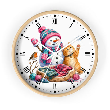 A round wall clock with a light wood frame and a white face featuring a winter illustration of a snowman in a pink knit hat and scarf, an orange tabby cat, a small bird, and colorful yarn balls.