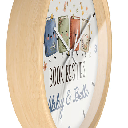A round wall clock with a light natural wood frame and a white face featuring four cute illustrated books with faces and arms, plus the text 'BOOK BESTIES' and 'Abby & Bella'.