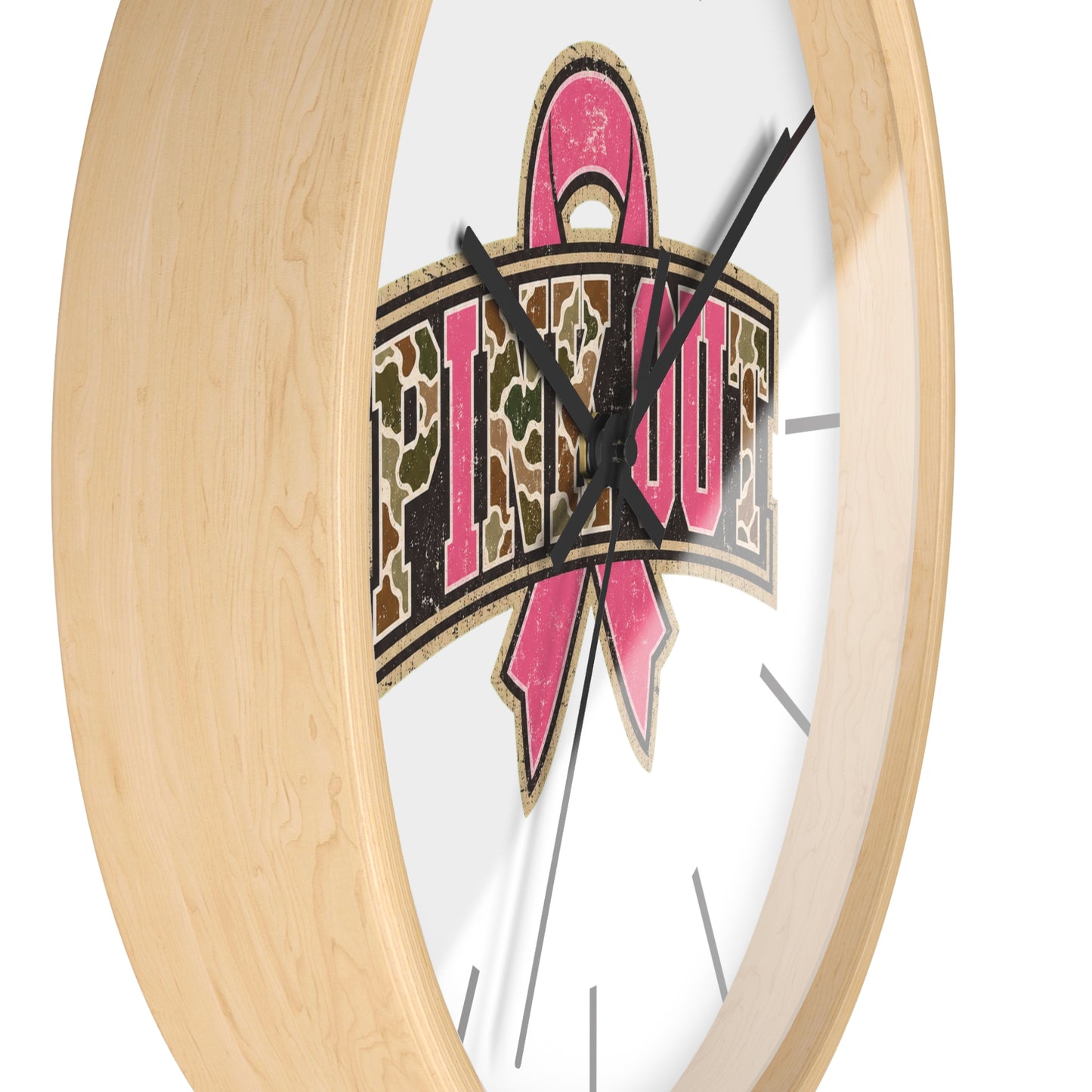 A close-up of a round wall clock with a light natural wood frame and a white face. The center features a graphic with a pink ribbon and the word 'PINK OUT' in a camouflage pattern.