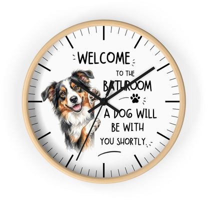 A round wall clock with a light wood frame and a white face, featuring a cartoon dog Australian shepherd illustration and the text 'WELCOME TO THE BATHROOM A DOG WILL BE WITH YOU SHORTLY.' on a plain white background.