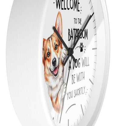 A round white wall clock with a humorous pet message printed on the face. The design features a watercolor illustration of a smiling corgi and the text 'WELCOME TO THE BATHROOM A DOG WILL BE WITH YOU SHORTLY'.