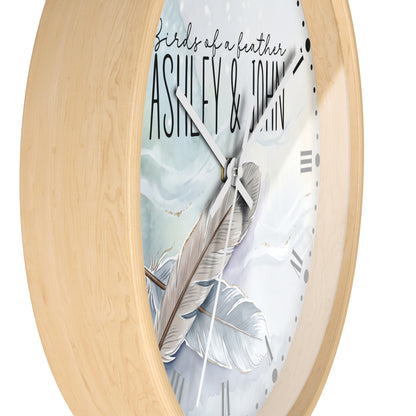 Oval wall clock with a light natural wood frame and a white face featuring a large feather illustration, black script text reading 'birds of a feather', and the names 'ASHLEY & JOHN'.
