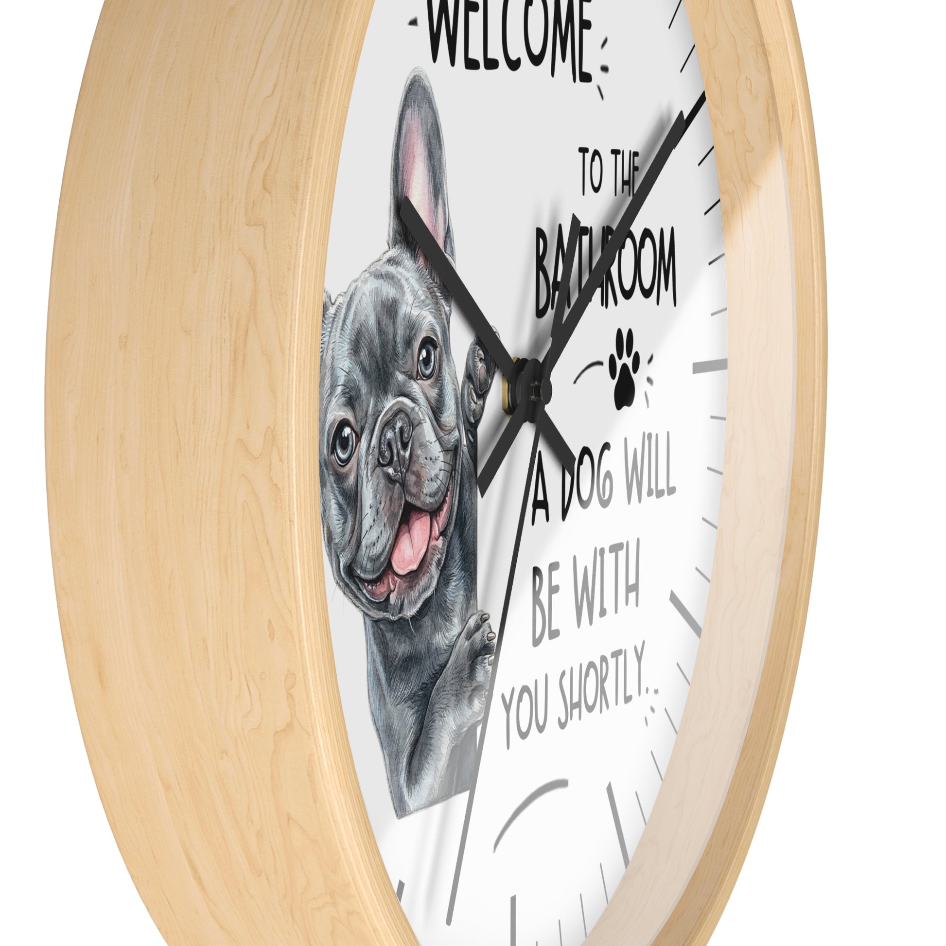 A round wall clock with a light natural wood frame and a white face featuring a black-and-gray French bulldog illustration and the text 'WELCOME TO THE BATHROOM A DOG WILL BE WITH YOU SHORTLY.