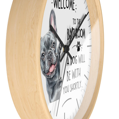 A round wall clock with a light natural wood frame and a white face featuring a black-and-gray French bulldog illustration and the text 'WELCOME TO THE BATHROOM A DOG WILL BE WITH YOU SHORTLY.