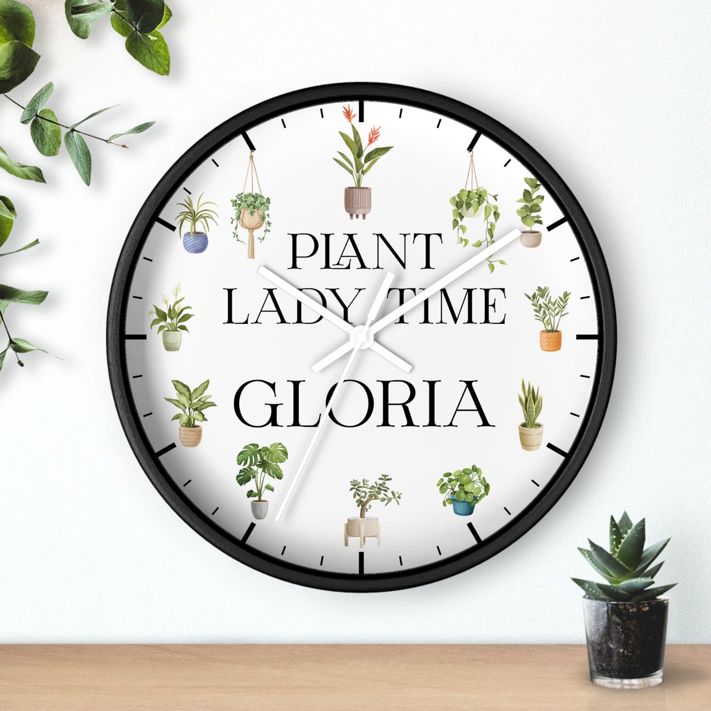 A round wall clock with a black frame and a white face, decorated with illustrations of potted plants and the text 'PLANT LADY TIME GLORIA'. The clock is shown hanging on a white wall above a wooden surface with small potted plants nearby.
