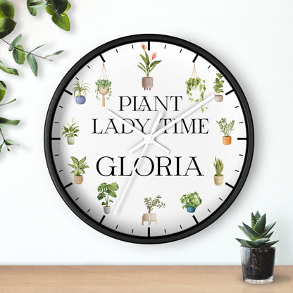 A round wall clock with a black frame and a white face, decorated with illustrations of potted plants and the text 'PLANT LADY TIME GLORIA'. The clock is shown hanging on a white wall above a wooden surface with small potted plants nearby.