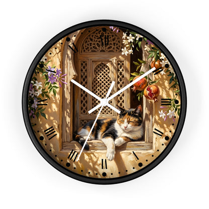 A round wall clock with a black frame and a detailed printed face showing a calico cat resting on a wooden window ledge, surrounded by flowers, pomegranates, and shadow patterns. The clock hands are white, and the background is a clean white studio setting.