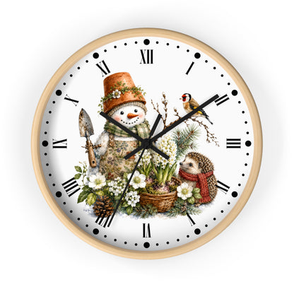 A round wall clock with a light natural wood frame and a white face featuring a detailed winter garden illustration. The design includes a snowman wearing a terracotta pot hat, a small bird, a hedgehog in a red scarf, white flowers, pinecones, and a garden spade, with black Roman numerals and hands.