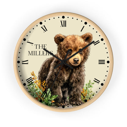 A round wall clock with a light wood frame and a cream-colored face, featuring an illustration of a brown bear cub standing among green plants and yellow flowers. The text 'THE MILLERS' is printed on the left side of the clock face, which is shown against a plain white background.