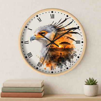 A round wall clock with a light wood frame hangs on a beige wall above a shelf with stacked books and a small potted succulent. The clock face features a watercolor-style illustration of a bird of prey over an African savanna sunset scene with an acacia tree.