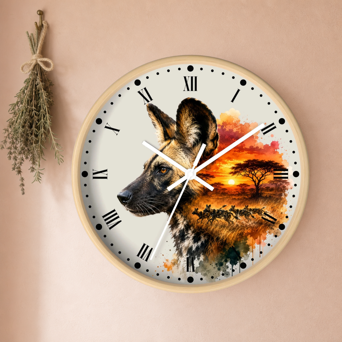 A round wall clock with a light wood frame, featuring a printed design of a wild dog profile on the left and a vibrant African savanna sunset on the right. The clock is mounted on a warm beige wall with a dried herb sprig hanging on the left.