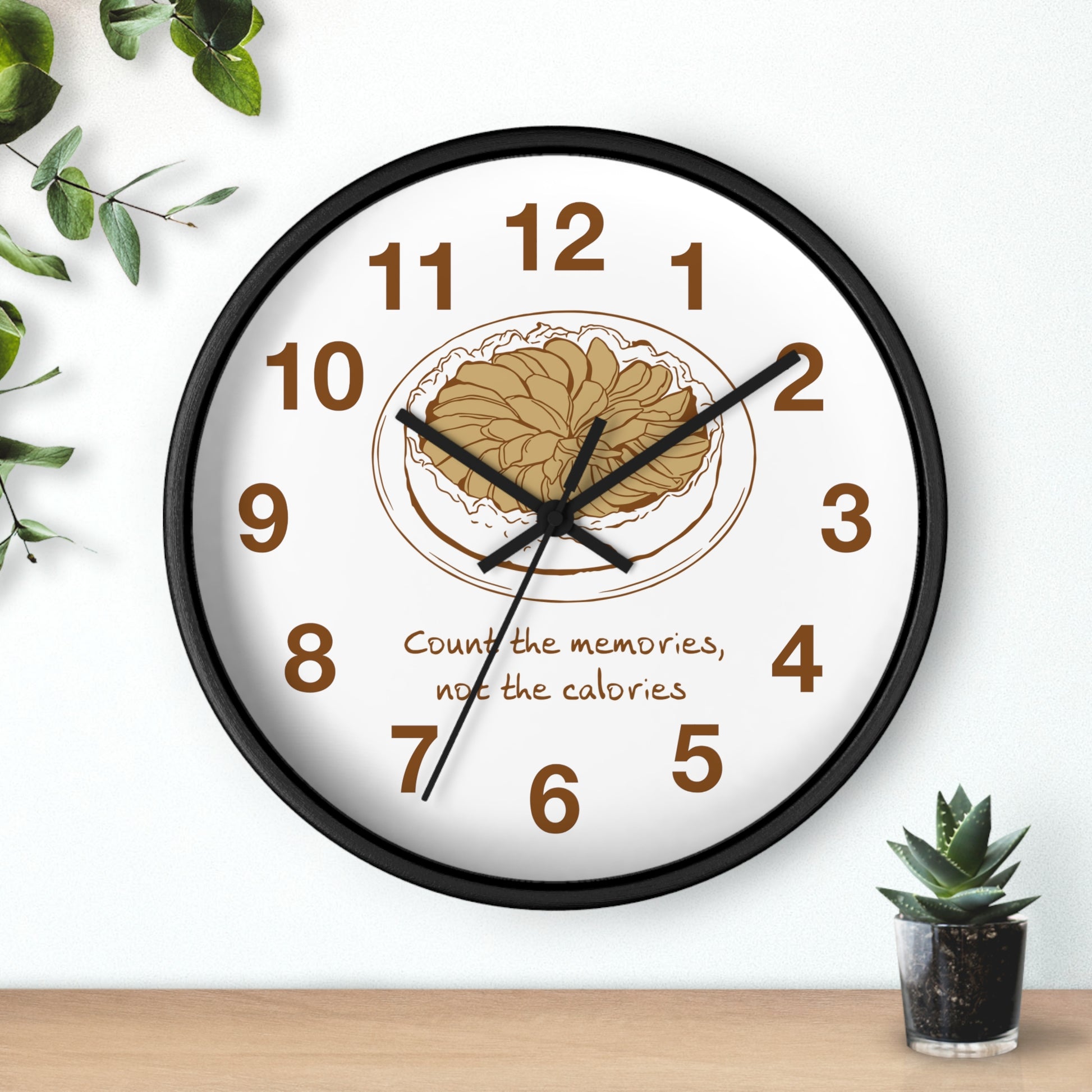 A round wall clock with a black frame and white face, featuring brown numerals, black hands, and a central illustration of a plate of pasta with the text 'Count the memories, not the calories.' The clock is shown hanging on a white wall above a wooden surface, with small green plants as decorative props.