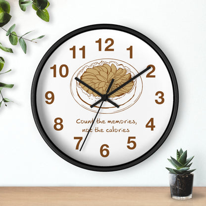 A round wall clock with a black frame and white face, featuring brown numerals, black hands, and a central illustration of a plate of pasta with the text 'Count the memories, not the calories.' The clock is shown hanging on a white wall above a wooden surface, with small green plants as decorative props.