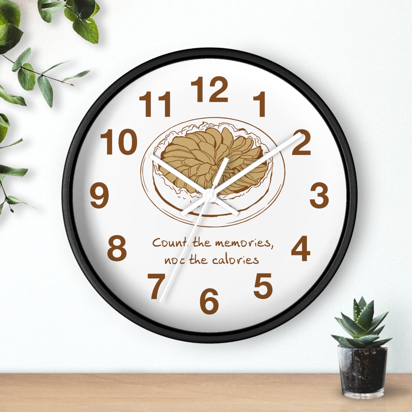 A round wall clock with a black frame and a white face, featuring a brown illustration of a plate of food at the center, brown numbers, and the text 'Count the memories, not the calories' below. The clock is shown hanging on a white wall above a wooden surface with small green plants in the corners.