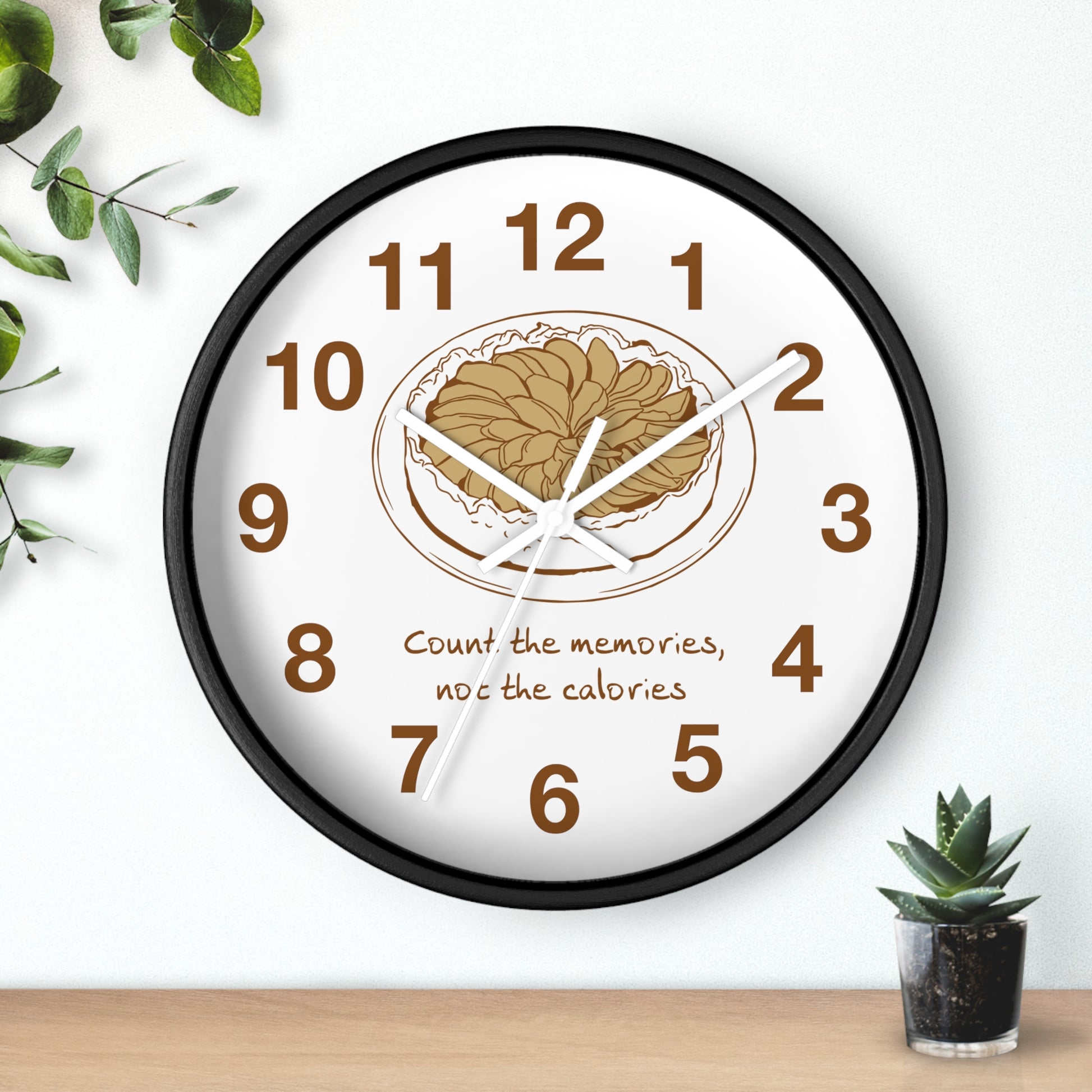 A round wall clock with a black frame and a white face, featuring a brown illustration of a plate of food at the center, brown numbers, and the text 'Count the memories, not the calories' below. The clock is shown hanging on a white wall above a wooden surface with small green plants in the corners.