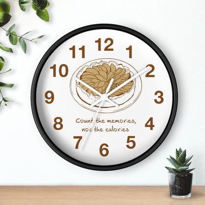 A round wall clock with a black frame and a white face, featuring a brown illustration of a plate of food at the center, brown numbers, and the text 'Count the memories, not the calories' below. The clock is shown hanging on a white wall above a wooden surface with small green plants in the corners.