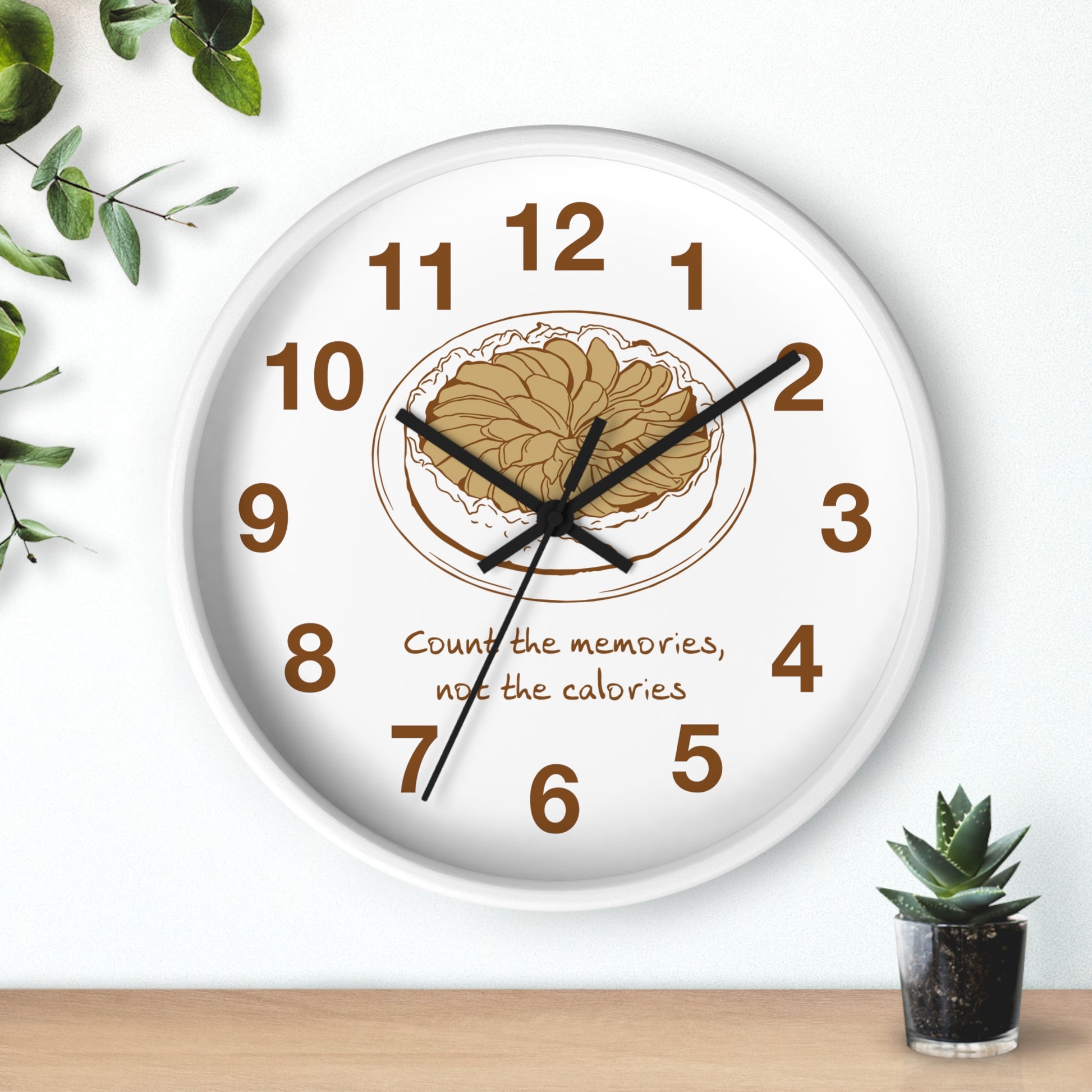 A round white wall clock with black hands and brown numerals, featuring a central illustration of a plate of pasta and the text 'Count the memories, not the calories'. The clock is shown hanging on a white wall above a light wood surface, with decorative green leaves and a small succulent plant nearby.