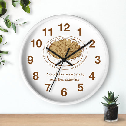 A round white wall clock with black hands and brown numerals, featuring a central illustration of a plate of pasta and the text 'Count the memories, not the calories'. The clock is shown hanging on a white wall above a light wood surface, with decorative green leaves and a small succulent plant nearby.
