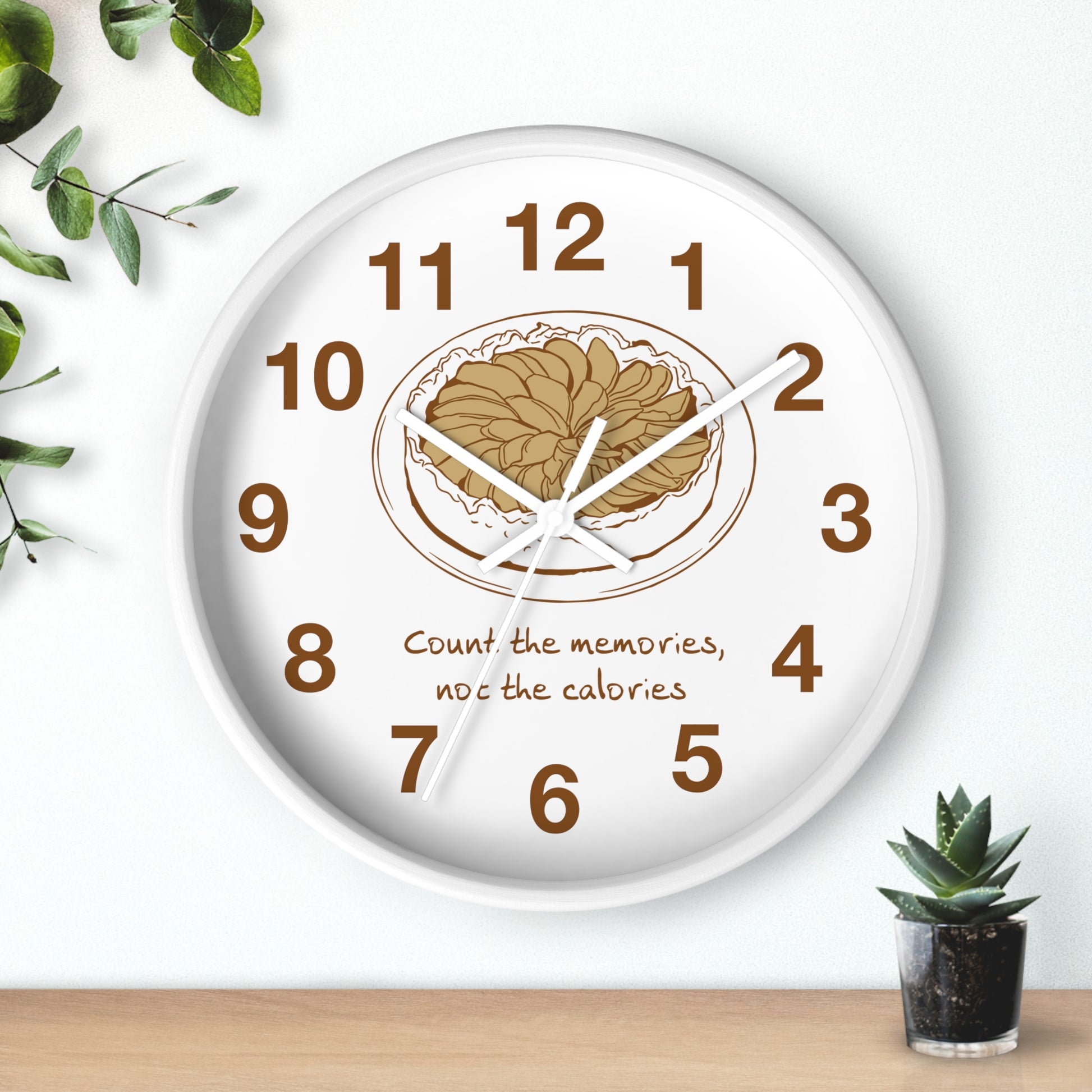 A round white wall clock with brown numerals and a brown plate of pizza graphic in the center, featuring the text 'Count the memories, not the calories' on a light wall above a wooden surface with small green plants.
