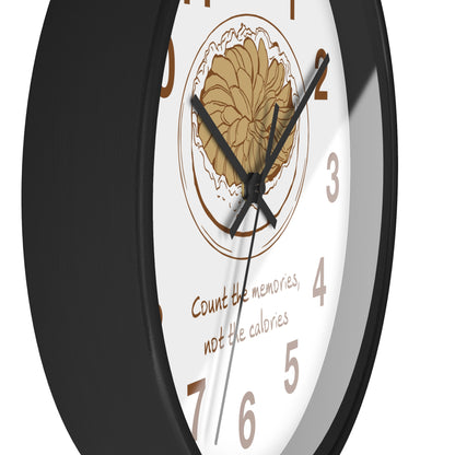A close-up of a round wall clock with a black frame and white face. The center features a tan floral illustration inside a circular border, with the text 'Count the memories, not the calories' in brown script below.
