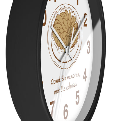 A close-up of a round wall clock with a black frame and a white face. The center features a tan floral illustration, brown Arabic numerals, silver hands, and the phrase 'Count the memories, not the calories' in brown script.