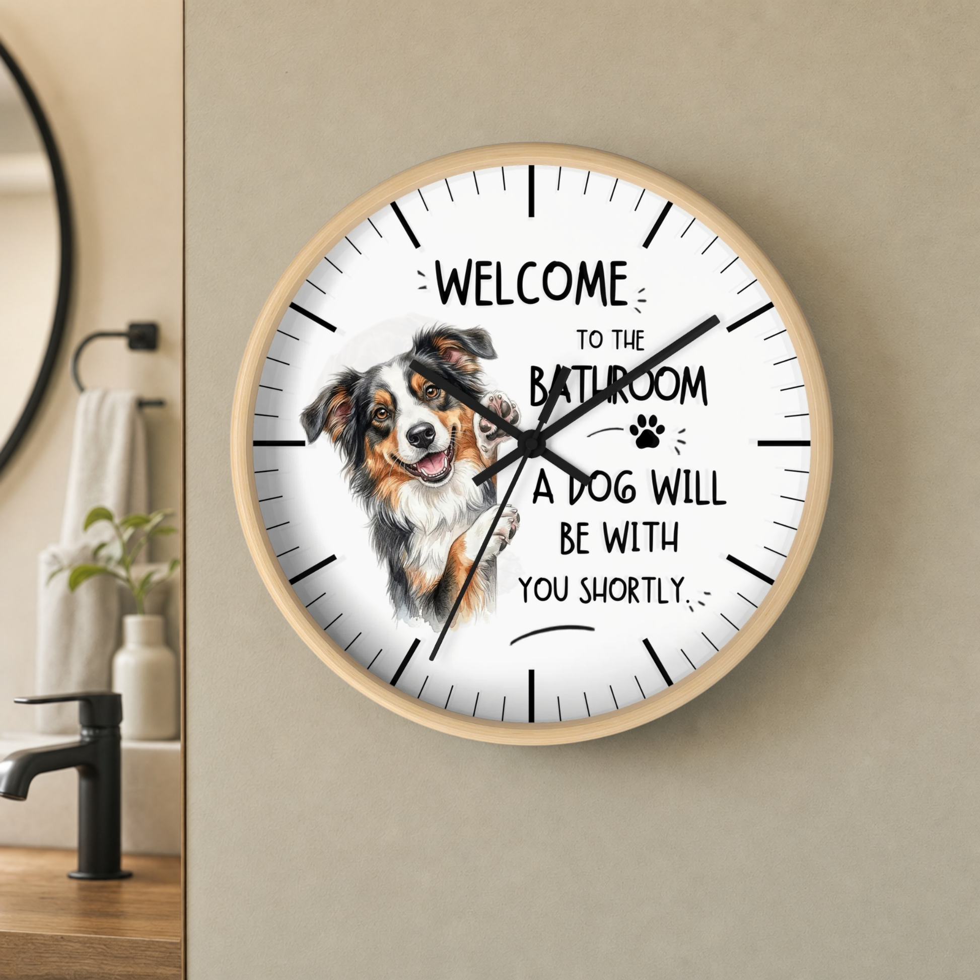 A round wall clock with a light wood frame hangs on a beige wall in a bathroom setting. The clock face is white with black text reading 'WELCOME TO THE BATHROOM A DOG WILL BE WITH YOU SHORTLY' and a watercolor illustration of a brown, white, and black dog.