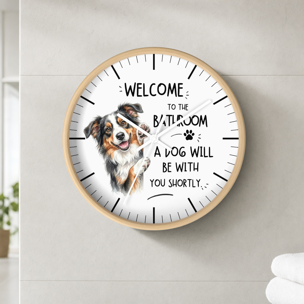 A round wall clock with a light wood frame and a white face, mounted on a light gray tiled wall. The clock features a watercolor illustration of a brown and white dog next to the text 'WELCOME TO THE BATHROOM A DOG WILL BE WITH YOU SHORTLY' with a small paw print.