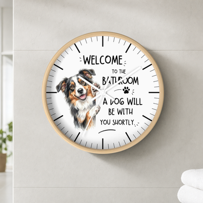 A round wall clock with a light wood frame and a white face, mounted on a light gray tiled wall. The clock features a watercolor illustration of a brown and white dog next to the text 'WELCOME TO THE BATHROOM A DOG WILL BE WITH YOU SHORTLY' with a small paw print.
