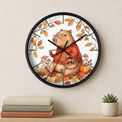 A round wall clock with a black frame and a whimsical autumn capybara illustration, hanging on a light beige wall above a shelf with books and a small succulent.